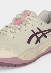 Ανοιχτό μπεζ αθλητικό παπούτσι ASICS με μωβ λογότυπο, ροζ λεπτομέρειες, λευκά κορδόνια και υφή από διχτυωτό υλικό, εμφανίζεται από πλάγια γωνία.