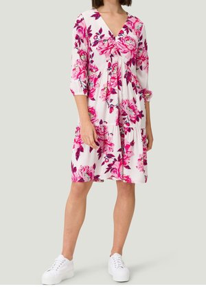 Femme portant une robe blanche jusqu'aux genoux avec des motifs floraux roses, des manches trois-quarts et des baskets blanches, debout devant un fond uni.