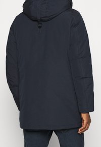 Tmavě modrá prošívaná bunda s konstrukční kapucí, vybavená zapínáním na přezky, dlouhými rukávy a rovným střihem. Hladká textura bez viditelných vzorů.