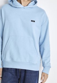 Felpa leggera color azzurro realizzata in tessuto morbido, con tasca a marsupio, polsini e orlo a costine, e un piccolo etichetta nera con logo sul petto.