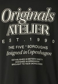 Μαύρο βαμβακερό μπλουζάκι με έντονο λευκό γραφικό κείμενο. Περιλαμβάνει "Originals ATELIER", "EST. 1990" και "Σχεδιασμένο στην Κοπεγχάγη."