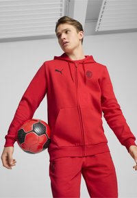 Rød zip-up hoodie med et sort logo og matchende sweatpants. Modellen holder en rød og sort fodbold. Glat stof, afslappet design.
