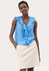 Haut sans manches bleu avec un décolleté en V à volants et un design fluide, associé à une jupe moulante de couleur crème. Texture du tissu lisse évidente.