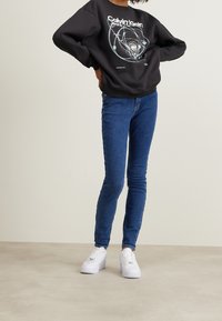 Svart sweatshirt med en cirkulär grafikdesign, kombinerad med blå skinny jeans och vita sneakers.