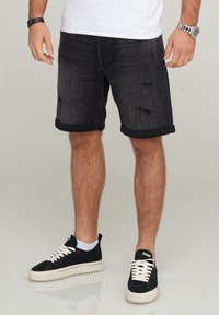 Pantalones cortos de mezclilla negros con puños enrollados, que presentan detalles desgastados. Combinados con zapatillas negras y calcetines blancos hasta los tobillos. Diseño simple y casual.