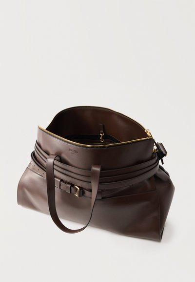 Τσάντα tote από σκούρο καφέ δέρμα με ανοιχτό φερμουάρ στην κορυφή, ρυθμιζόμενο λουρί και δύο λουριά ώμου σε ουδέτερο φόντο.
