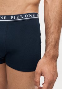 Muž nosící tmavě modré boxerky s páskem značky Pier One, zaměřeno na spodní část trupu a pravou ruku.