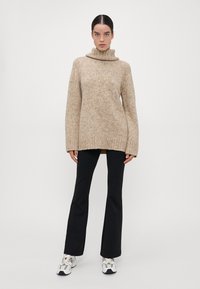 Beige oversized gebreide trui met hoge kraag, geribbelde zoom en lange mouwen, gecombineerd met zwarte flare broeken en witte sneakers.