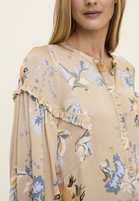 Blouse beige avec des motifs floraux en bleu, jaune et vert. Présente des accents froncés aux épaules, une patte de boutonnage et un tissu texturé.