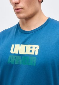 Νέος άνδρας που φοράει μπλε μπλουζάκι με στρογγυλή λαιμόκοψη με τις λέξεις "UNDER ARMOUR" τυπωμένες με μεγάλα γράμματα στο στήθος.