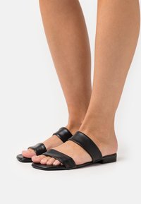 Fötter i svarta platta sandaler med två breda läderremmar, en vid tårna och en över mitten av foten, på en vit bakgrund.