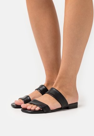 Pieds portant des sandales plates noires avec deux larges contreforts en cuir, l'un près des orteils et l'autre sur le milieu du pied, sur un fond blanc.