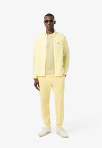 Uomo che indossa una camicia a righe gialle con bottoni sopra una t-shirt gialla, pantaloni felpati gialli, scarpe bianche e grandi occhiali da sole marroni su sfondo bianco.