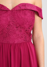 Fuchsia-klänning med en avskuren axelspets överkropp som har blommönster och en plisserad kjol, tillverkad av lätt tyg.