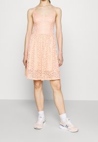 Robe en dentelle pêche avec bretelles réglables, corsage ajusté, jupe évasée et motif floral. Portée avec des baskets de sport rose clair.