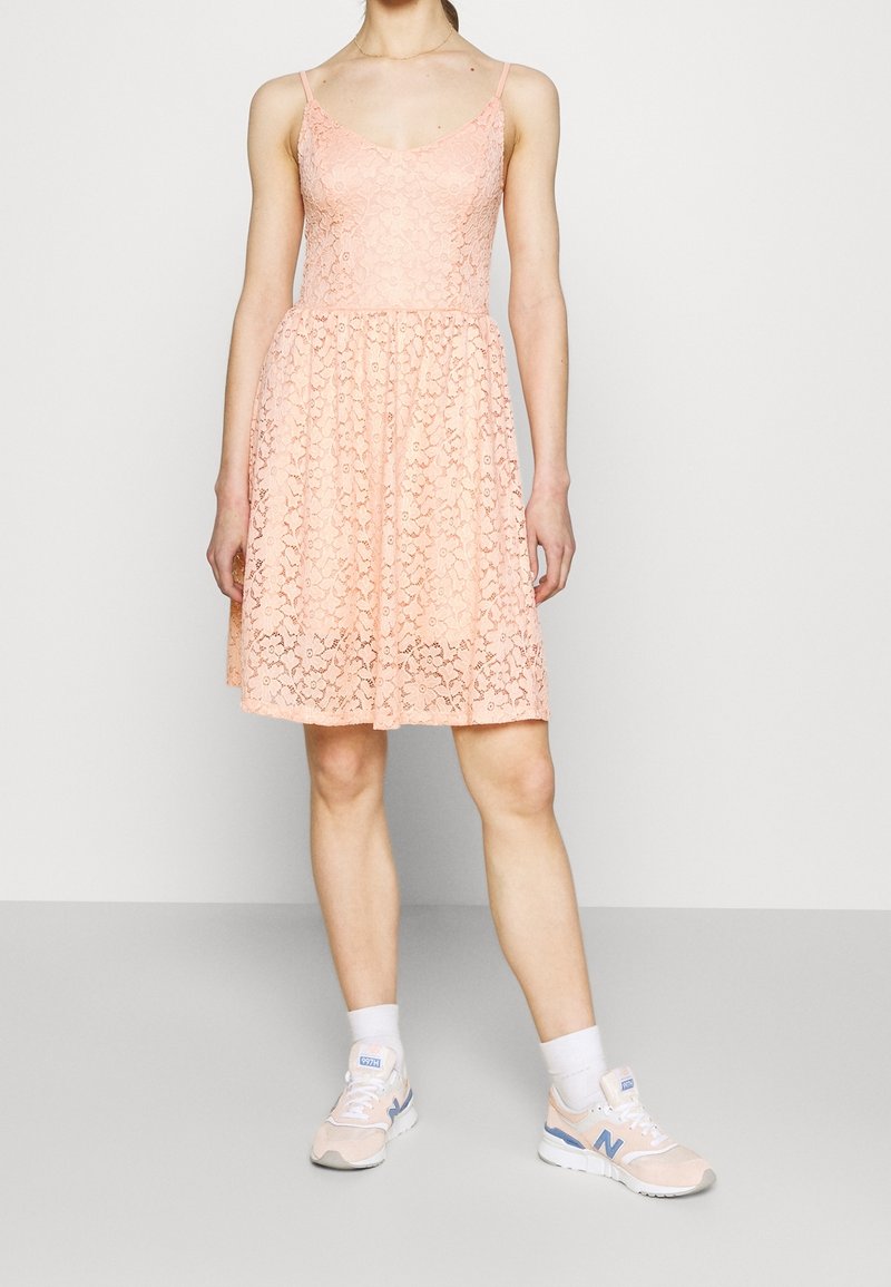 Robe en dentelle pêche avec bretelles réglables, corsage ajusté, jupe évasée et motif floral. Portée avec des baskets de sport rose clair.