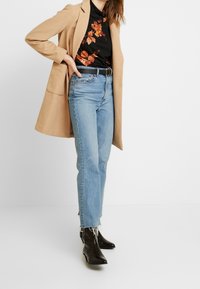 Beżowy długi płaszcz na czarnym kwiatowym topie, w połączeniu z jasnoniebieskimi spodniami o wysokim stanie i czarnymi botkami za kostkę z ozdobnymi przeszyciami.