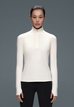 Une femme porte un haut blanc ajusté à col montant et à manches longues, avec des poignets à passe-pouces, ainsi qu’un legging noir, sur fond gris.