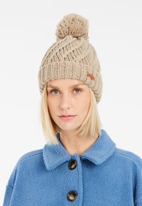 Berretto beige lavorato a maglia con un grande pompon sopra, caratterizzato da un motivo testurizzato e un piccolo etichetta in pelle. Realizzato in filato spesso e morbido.
