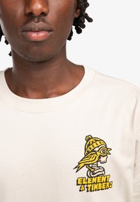 Camiseta de manga corta color crema con un gráfico amarillo y negro de un pájaro sobre una calavera y el texto "ELEMENT & TIMBER!" en el pecho.