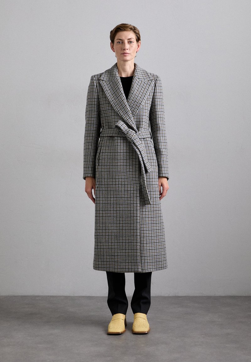 Long, checkered coat in grey and black with a belted waist, paired with black pants and yellow woven shoes, standing against a neutral background.