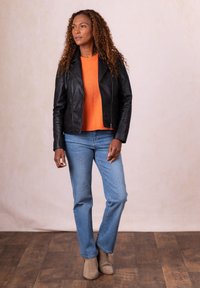 Black leather jacket, orange sweater, light blue jeans, and beige ankle boots. Curly hair, standing on wooden floor against a neutral backdrop.