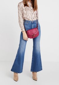 Borsa a tracolla rossa in pelle con pattina e accessori dorati, indossata con una blusa a fantasia e jeans a palazzo blu. Stivali beige con tacco completano l'outfit.