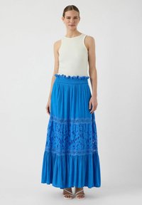Een blauwe maxirok met kantdetails en een gerimpelde zoom, gecombineerd met een mouwloze witte top van ribbelstof.