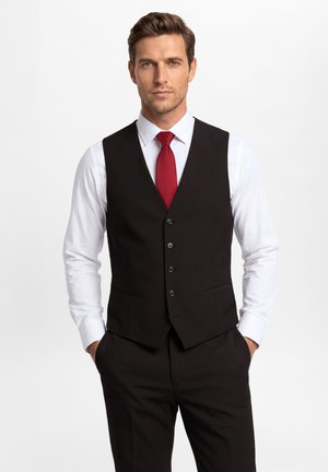 Mężczyzna w czarnej kamizelce garniturowej, białej koszuli i czerwonym krawacie stoi z rękami w kieszeniach na jednolitym białym tle.