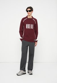 Camisa de manga longa bordeaux com o texto branco "NIKE AIR", calças largas cinzas com fecho lateral e ténis cinzas com detalhes pretos. Óculos de sol pretos.
