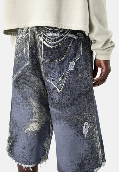 Personne portant un short en jean ample et usé aux bords effilochés, ainsi qu'une chemise à manches longues beige clair recouvrant partiellement la ceinture.