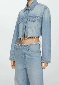 Chaqueta corta de mezclilla azul claro con dobladillo desflecado, cierre de botones en la parte delantera y dos bolsillos en el pecho, combinada con jeans a juego de corte recto.