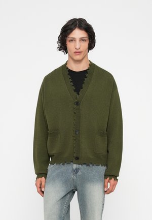 Jonge man met krullend donker haar, gekleed in een olijfgroene knoopcardigan met rafelige randen en lichtblauwe versleten spijkerbroek.