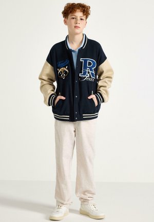 Ragazzo dai capelli rossi che indossa una giacca varsity blu navy e beige con una toppa animale, polo azzurro chiaro, pantaloni beige e sneakers bianche, in piedi con le mani nelle tasche.