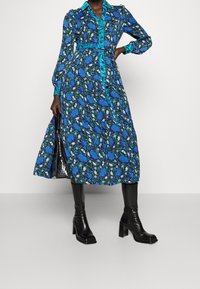 Abito lungo blu floreale con vita aderente, chiusura con bottoni sul davanti e colletto teal a contrasto. Abbinato a stivali neri con tacco al ginocchio.