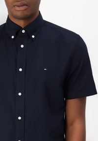 Camicia a maniche corte blu navy con bottoni in cotone, con colletto e bottoni bianchi. Presenta un piccolo logo rosso, bianco e blu sul petto.