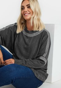 Grå långärmad tröja med ribbad textur, med silverbesatta detaljer längs axlarna. Kombinerad med blå denimjeans.