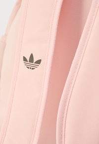 Rožinės spalvos kuprinė su tekstūruota medžiaga, tamsiai pilku Adidas logotipu ir kontrastinėmis šviesiai rožinėmis petnešomis. Paprastas dizainas, be papildomų raštų.