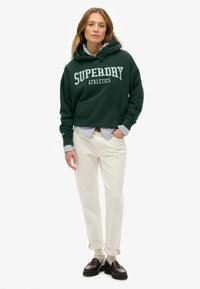 Mujer con sudadera oscura verde "Superdry Athletics", camiseta a rayas, pantalones crema, calcetines beige y mocasines marrones, de pie con la mano en el bolsillo.