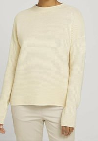 Krämfarbener, gerippter Strickpullover mit lässigem Schnitt, Rundhalsausschnitt und langen Ärmeln, kombiniert mit hellbeigen Hosen.