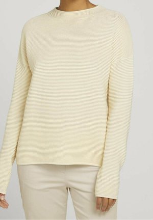 Krämfarbener, gerippter Strickpullover mit lässigem Schnitt, Rundhalsausschnitt und langen Ärmeln, kombiniert mit hellbeigen Hosen.