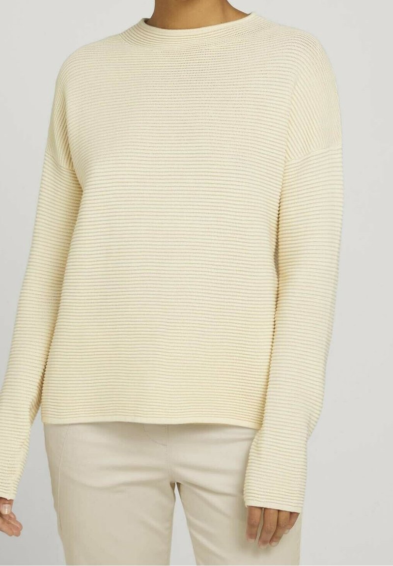 Krämfarbener, gerippter Strickpullover mit lässigem Schnitt, Rundhalsausschnitt und langen Ärmeln, kombiniert mit hellbeigen Hosen.