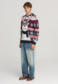 Maglione di Natale lavorato a maglia con design di renna in bianco, rosso e blu navy, decorato con fiocchi di neve e righe. Abbinato a jeans azzurri e scarpe marroni.