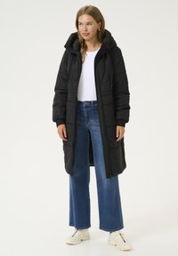Manteau doudoune noir avec capuche, doté d'une fermeture éclair et de poignets élastiques, associé à un jeans bleu à jambes larges et des baskets blanches.