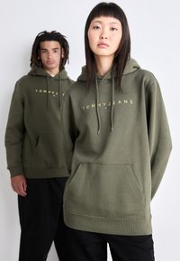 Olíva zöld kapucnis pulóver, elülső zsebbel, húzózsinórral és arany "TOMMY JEANS" felirattal a mellkason.