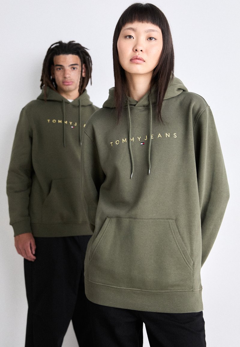 Felpa con cappuccio verde oliva, dotata di tasca frontale, dettagli con cordino e scritta dorata "TOMMY JEANS" sulla parte anteriore.