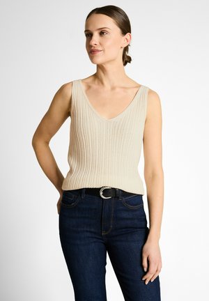 Frau mit braunen Haaren zu einem niedrigen Dutt gebunden, trägt ein cremefarbenes geripptes ärmelloses V-Ausschnitt-Top und dunkelblaue Jeans mit einem schwarzen Gürtel.