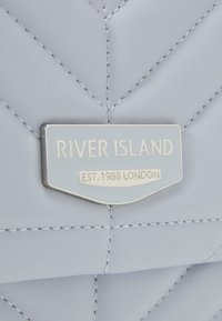 Błękitna torebka pikowana z srebrną tabliczką z logo, na której widnieje napis "RIVER ISLAND EST. 1988 LONDON." Gładka tekstura z wyraźnymi przeszyciami.