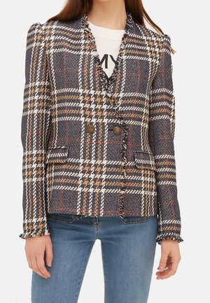 Vrouw draagt een dubbelgeknoopte geruite blazer met franjes aan de randen over een wit overhemd en blauwe spijkerbroek.