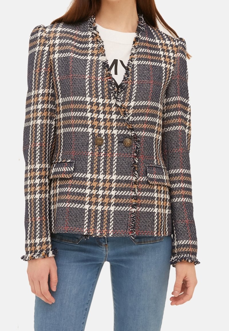 Femme portant un blazer à carreaux croisé avec des bords frangés sur une chemise blanche et un jean bleu.